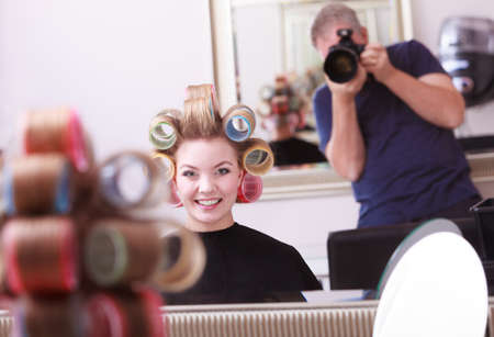 Portrait of happy woman in beauty salon. Cheerful blond girl with hair curlers rollers by hairdresser. Hairstyle.の写真素材