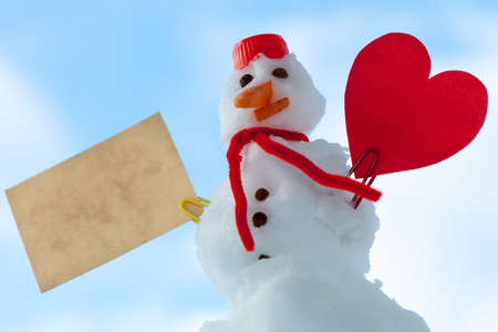 Little happy christmas snowman in red scarf and with clip heart love symbol valentine paper card text message outdoor. Winter season seasonal specific valentine's day. Blue sky background.の写真素材