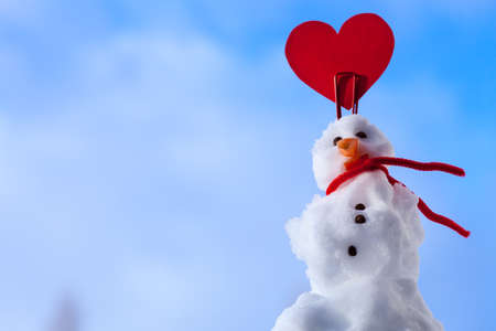 Little happy valentine snowman in red scarf and with clip red heart love symbol outdoor. Winter season seasonal specific valentine's day. Blue sky background.の写真素材