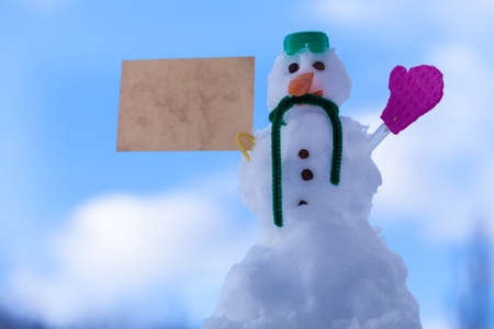 Little happy christmas snowman in green scarf and with paper card message copyspace copy space outdoor. Winter season seasonal specific valentine's day. Blue sky background.の写真素材