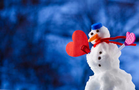 Little happy christmas snowman in blue screw top as hat red scarf and with heart love symbol outdoor. Winter season seasonal specific valentines day. Blurred background with trees.の写真素材
