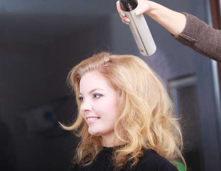 Girl with blond wavy hair by hairdresser  Hairstylist with hairspray and female client  Young woman...