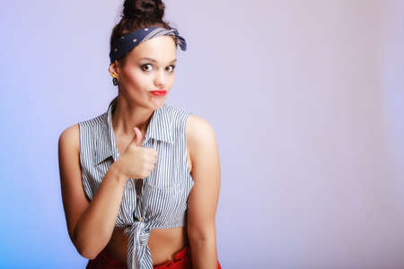 Portrait of fashionable pin-up girl young woman with bun showing thumb up hand sign gesture. Blank copy space on violet background. Success. Studio shot.の写真素材