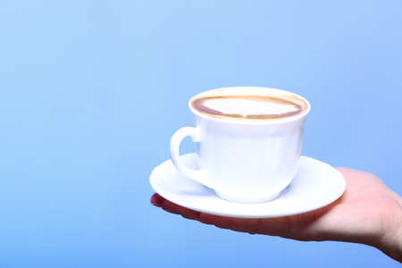 Closeup female hand holding a cup of latte coffee cappuccino hot drink in autumn winter time copyspace blue backgroundの写真素材