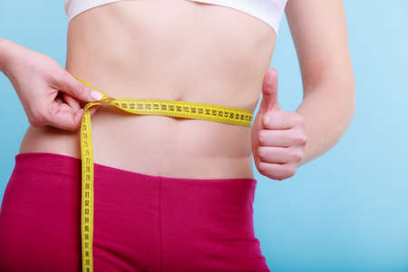 Time for diet slimming weight loss. Health care and healthy nutrition. Young fitness woman fit girl with measure tape measuring her waistline showing thumb up success hand gesture on blue greenの写真素材