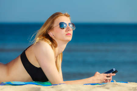 Summer vacation. Sexy girl in bikini sunbathing tanning on the beach. Young woman relaxing with mobile phone on the sea coast. Summertime.の写真素材