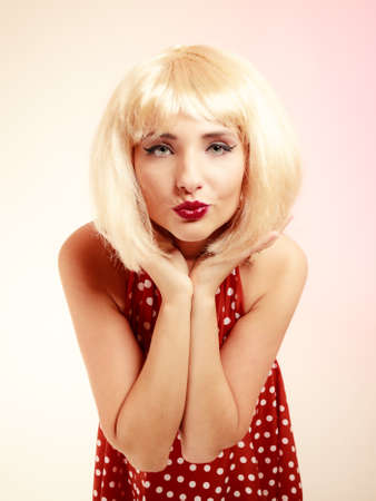 Vintage pinup style. Portrait of stylish woman blowing a kiss on pink. Girl in blond wig and retro spotted red dress. Disguise.の写真素材
