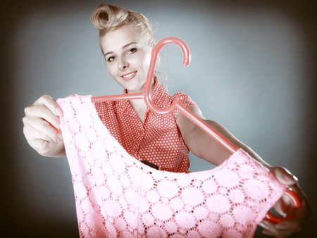 Pinup blond girl young woman in retro style buying clothes. Client customer holding pink dress on gray. Retail and sale. Studio shot.の写真素材