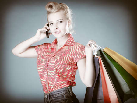 Pinup girl young woman in retro style buying clothes calling on the mobile phone. Client holding shopping bags on gray. Retail and sale.の写真素材