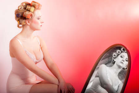 Sexy thoughtful woman preparing to party, girl in underwear with curlers in hair looking at the mirror black white reflection pink backgroundの写真素材