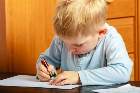 Happy childhood. Blonde boy child kid with pen writing drawing on paper doing homework. At home.の写真素材