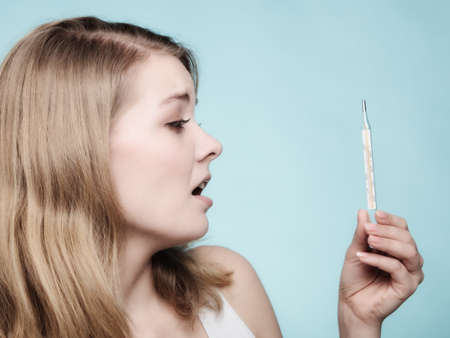 Flu cold grippe. Woman having high temperature. Sick girl with fever checking mercury thermometer on blue.の写真素材