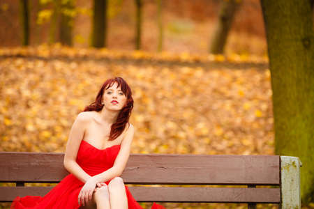 Beautiful young woman in elegant red dress sitting on bench. Girl relaxing on autumnal park. Fall leisure. Outside.の写真素材