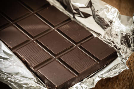 Sweet food. Dark chocolate bar in opened silver foil wrapping on wooden table.の写真素材