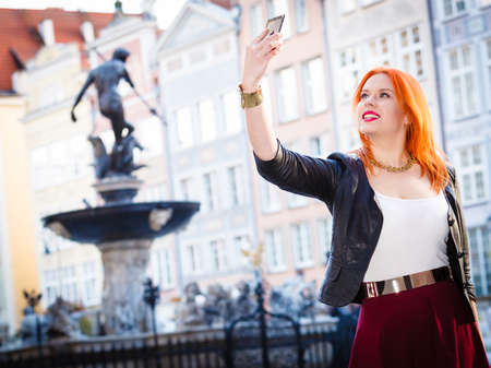 Traveler woman redhair fashion girl taking self picture selfie with smartphone camera outdoors in european city old town Gdansk. Neptune fountain in the background Polandの写真素材