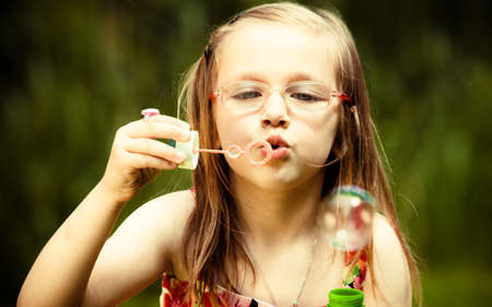 Children happiness and carefree concept. Little girl having fun blowing soap bubbles in park green blurred backgroundの写真素材