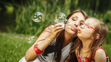 Family happiness and carefree concept. Mother and daughter little girl having fun blowing soap bubbles together in park, green blurred backgroundの写真素材
