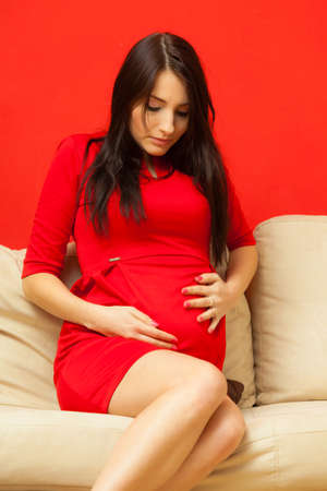 Pregnancy, motherhood and happiness concept. Beautiful sexy stylish elegant pregnant woman in red dress relaxing on sofa and touching her bellyの写真素材