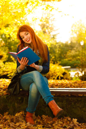 Fall lifestyle concept. Redhaired young woman girl relaxing in autumn park reading book, sitting...
