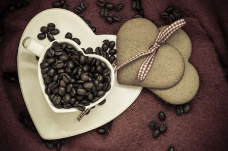 Coffee love concept. Coffee beans in heart shaped white cup and sweet cookie gingerbread with ribbon on brown cloth background. Top viewの写真素材