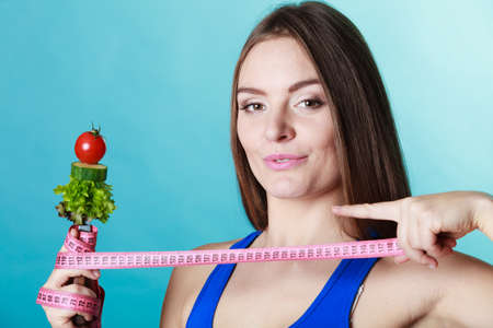 Dieting weight loss concept. Sporty girl fitness woman holding fork with fresh mixed vegetables and measuring tape on blue backgroundの写真素材