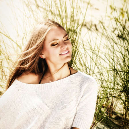 Portrait of beautiful young woman breathing fresh air with closed eyes while sitting on beach sand...
