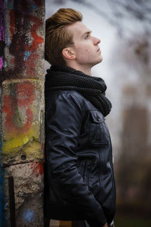Face profile thoughtful trendy man outdoor in city setting, male model wearing winter clothes black jacket and scarf against colorful graffiti wallの写真素材