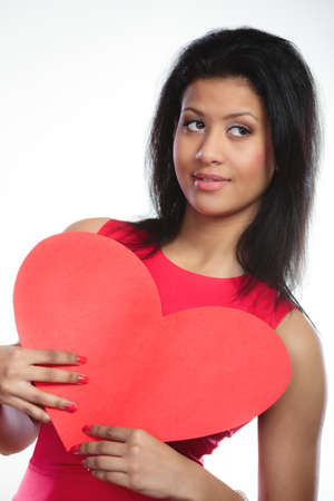 Happiness and valentines day concept. Fashion young woman holding red heart card. Girl mixed race african caucasian in fashionable red dress posing with love symbol on grayの写真素材