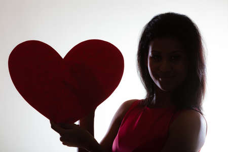 Happiness and valentines day concept. Silhouette of woman with red heart card. Girl mixed race african caucasian in fashionable red dress posing with love symbol on grayの写真素材