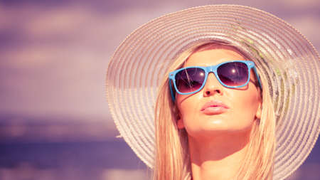 Fashion, happiness and lifestyle concept. Lovely blonde girl in hat and color sunglasses walking on beach. Young woman relaxing on the sea coast.の写真素材