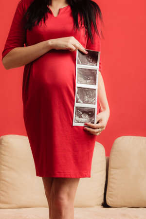 New life concept. Pregnancy, motherhood and happiness. Closeup tummy of pregnant woman wearing elegant red dress, ultrasound scan in hand indoorの写真素材