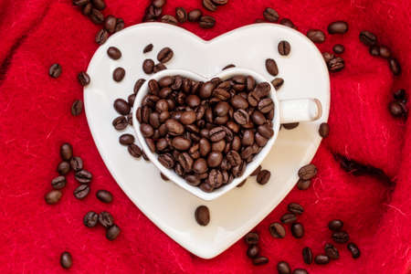 Coffee klatsch java concept. Heart shaped white cup filled with roasted coffee beans on red cloth...