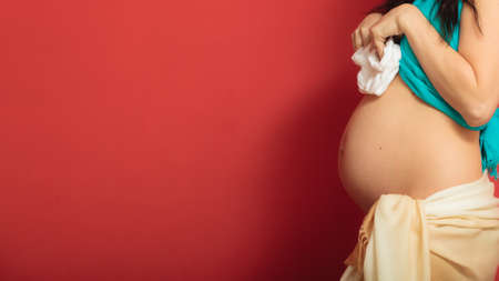 Pregnancy, motherhood and happiness concept. pregnant woman holding small shoes boots for the unborn baby on red backgroundの写真素材