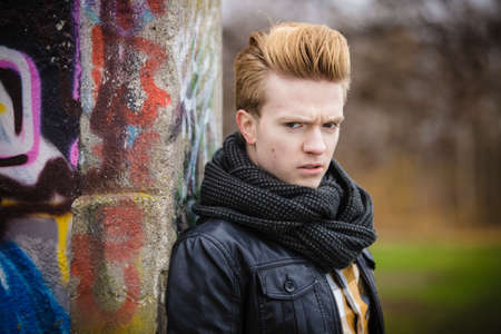Portrait of handsome trendy man outdoor in city setting, male model wearing winter clothes black jacket and scarf against colorful graffiti wallの写真素材