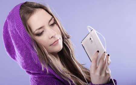 young woman with smart phone listening music. Teen stylish long hair girl in hood relaxing or learning language. Studio shot on violet.の写真素材