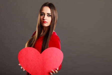 Valentines day concept. Brunette long hair young woman in red dress holding big heart love symbol dark gray backgroundの写真素材