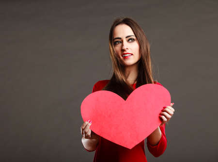 Valentines day love and relationships concept. Brunette woman long hair girl in red outfit holding heart love symbol, text area dark gray backgroundの写真素材