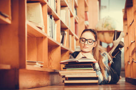 Clever female student girl in glasses lying on floor in college library with stack books readingの写真素材