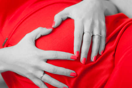 Pregnancy, motherhood and happiness concept. stylish elegant pregnant woman in red dress relaxing on sofa, making heart shape with hands over belly, symbol of loveの写真素材