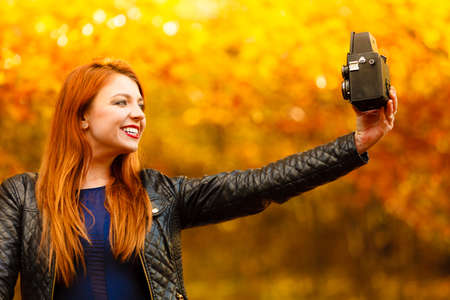 fashionable redhair tourist girl young woman taking self photo picture with old vintage camera. Beauty autumn park, golden leaves backgroundの写真素材