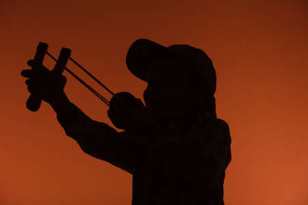 Children upbringing problems. Kid holding slingshot in hands. Bad naughty boy silhouette shoots from a wooden sling on orange backgroundの写真素材