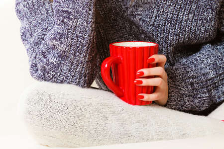 Hot beverage. Closeup female hand holding red cup mug hot drink tea or coffee, sitting on couch. Woman in warm sweater warming herselfの写真素材