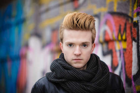 Portrait of handsome trendy man outdoor in city setting, male model wearing winter clothes black jacket and scarf against colorful graffiti wallの写真素材