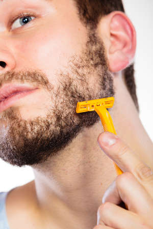 Health beauty and skin care concept. Closeup of male face. Young man guy styling beard holding disposable yellow razor blade white background.の写真素材