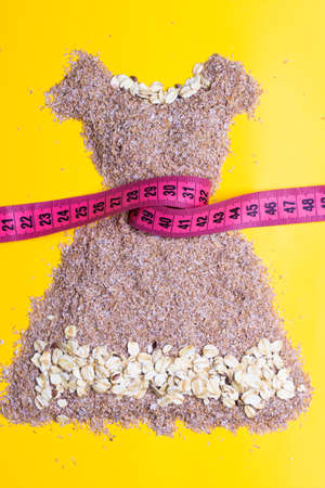 Dieting healthy eating slim down concept. Female dress shape made from cereal bran with measuring tape around thin waistline on yellowの写真素材
