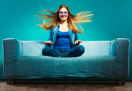 Fashion young woman in full length. Fashionable girl wearing denim sitting on couch wind in hair blue backgroundの写真素材