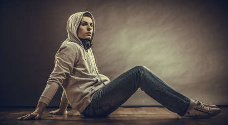 Music passion youth concept. Serious hooded man teen boy with headphones sitting daydreaming on floor grunge backgroundの写真素材