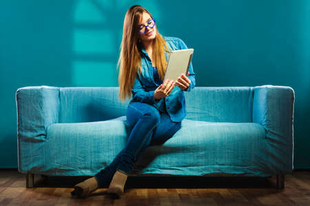 Modern technologies leisure and young people concept. fashionable woman wearing jeans with tablet sitting on couch blue colorの写真素材