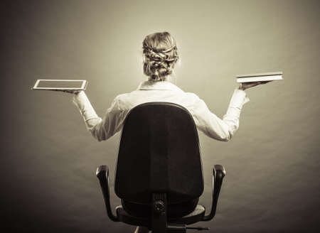 Ebook vs book. Woman sitting on chair holding traditional book and e-book reader tablet touchpad pc back view grunge background.の写真素材