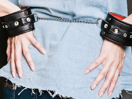 Arrest and jail. Closeup leather handcuffs on female hands, criminal woman prisoner girl back view on dark. Punishment.の写真素材